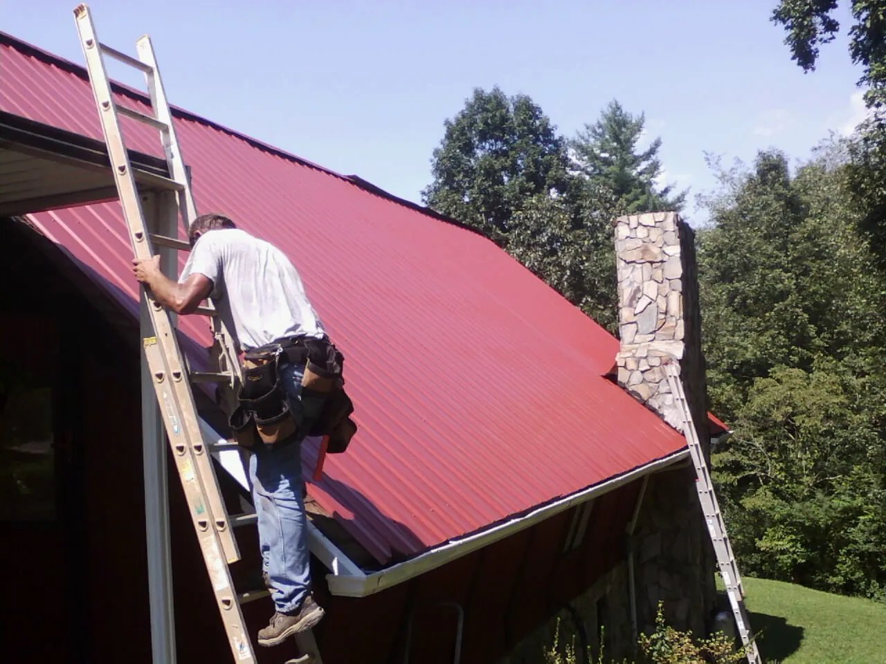 Roofer on a ladder beside a red metal roof and stone chimney — Soffit & Fascia Repair by Roofing Brothers in Cary